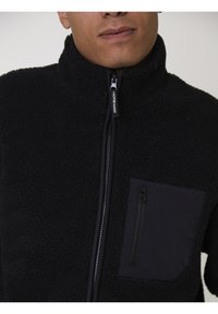 Uomo che indossa una giacca in pile nera con zip frontale e una tasca sul petto con zip, parzialmente visibile dal collo al petto.