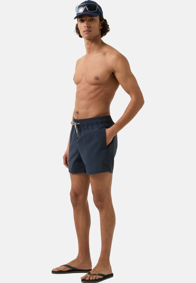 Marinefarbene Badehose mit elastischem Bund, Kordelzug, Seitentaschen und einem Logopatch. Getragen mit Zehenschuhen und einer Kappe.