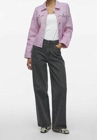 Chaqueta de mezclilla rosa claro, top blanco sin mangas, pantalones de mezclilla negros de tiro alto y pierna ancha, y zapatos con estampado. Vista frontal, manos en los bolsillos.