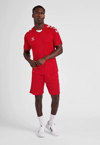 Rood atletisch shirt en korte broek, met witte accenten op de schouders en een logo op de borst. De stof lijkt glad en lichtgewicht. Witte sokken en schoenen.