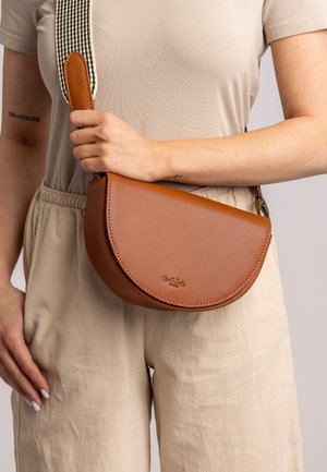 Persoon in beige kleren houdt een bruin leren halve maan schoudertas met een zwart-witte geweven strap over de borst.