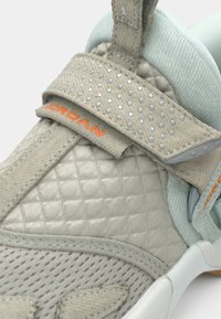 Gros plan sur la bride beige d'une chaussure Jordan avec de petits clous ronds, tissu matelassé, texte orange « JORDAN » et doublure intérieure rembourrée bleu clair.