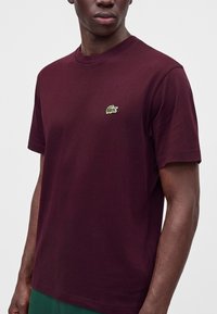 Homme portant un t-shirt uni bordeaux foncé à manches courtes avec un petit logo crocodile vert sur la poitrine gauche, sur fond blanc.
