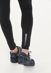 Svarta leggings tillverkade av stretchmaterial, med ett reflekterande randmönster på underbenet, matchade med mörka träningsskor.
