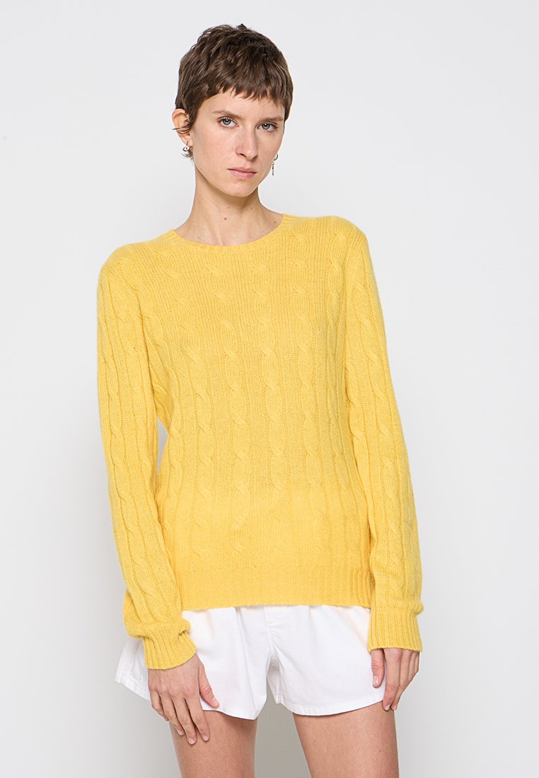 Kobieta z krótkimi włosami w żółtym swetrze w sploty i białych szortach, stojąca na jednolitym jasnym tle.