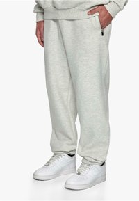 Light grey sweatpants with a soft texture, featuring a side zip pocket and embroidered branding. Paired with white sneakers.
