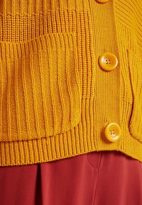 Pletený žlutý cardigan s žebrovanou strukturou, s velkou náprsní kapsou a třemi kulatými oranžovými knoflíky vpředu.