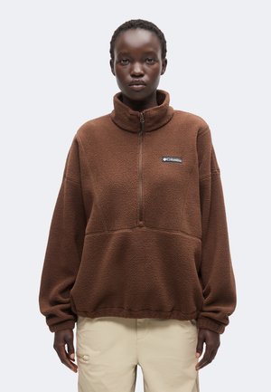 Person iført en brun Columbia fleece-pullover med halv lynlås og beige bukser, stående mod en ensfarvet lys baggrund.
