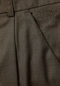 Brunt teksturert stoff på bukse med beltestropp, frontssøm og "HOUSE OF SUNNY" brodert over lommen.