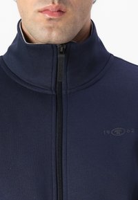 Chaqueta de color azul marino con cremallera y un alto cuello, que presenta una textura suave, un logo con "1962" en el lado izquierdo y una cremallera central.