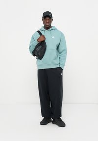 Hoodie azul claro com um bolso frontal e logótipo, combinado com calças largas pretas e sapatos pretos. A transportar uma mala à tiracolo preta.