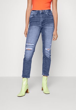 Vrouw draagt een high-waisted gescheurde blauwe spijkerbroek, neon groene enkellaarsjes met blokhakken en een oranje mouwloos topje.