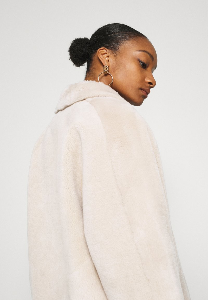 Manteau en fausse fourrure beige avec un large col, coupe ample et surface texturée. Le modèle montre son profil et porte des boucles d'oreilles en or.