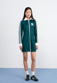En ung kvinna står mot en enkel bakgrund, klädd i en grön Adidas-klänning med vita ränder och svarta sandaler, med händerna i fickorna.