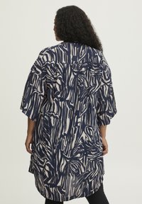 Lang, løstsiddende kimono med et navy og beige abstrakt mønster. Lavet af et letvægtsstof med brede ærmer og en let buet kant.
