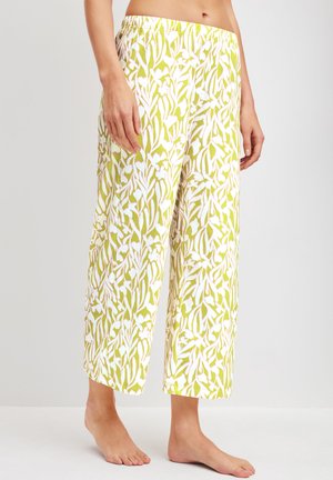 Femme portant un pantalon large taille mi-haute avec un motif floral vert et blanc, debout pieds nus devant un fond clair uni.