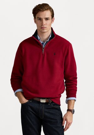 Sudadera de forro polar roja con cuello de media cremallera, que presenta un pequeño logo en el pecho, usada sobre una camisa azul clara y jeans oscuros.