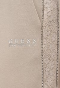 Μπεζ τοπ με το λογότυπο «GUESS LOS ANGELES» σε ασημί. Λεπτομέρεια με διακοσμητική δαντέλα και χάντρες κατά μήκος της μιας πλευράς. Απαλή, λεία υφή.