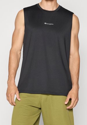 Man draagt een zwart mouwloos Champion-shirt en olijfgroene broek, staat met ontspannen armen langs het lichaam.