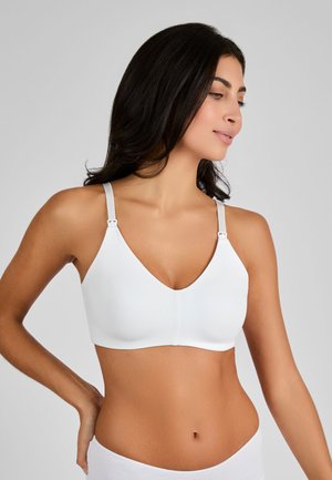 Vrouw draagt een witte naadloze voedingsbeha met verstelbare bandjes, kijkt naar de zijkant tegen een effen grijze achtergrond.