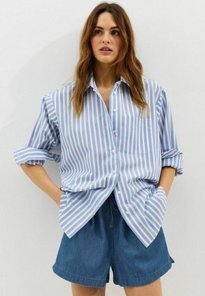 Donna che indossa una camicia con bottoni a righe blu e bianche con maniche arrotolate e pantaloncini di jeans blu, in piedi con le mani nelle tasche.