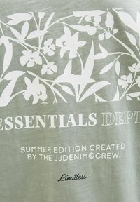 Gaiši zaļš audums ar baltu ziedu un lapu rakstu; teksts baltā un melnā krāsā, kas vēsta "ESSENTIALS DEPARTMENT" un "Limitless."
