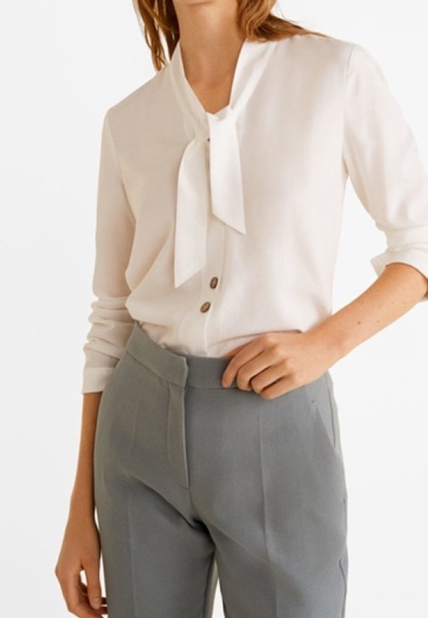 Femme portant un chemisier blanc avec un nœud au cou et un pantalon taille haute beige-gris sur mesure avec des boutons sur le devant du chemisier.