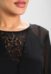 Donna che indossa un top nero con maniche trasparenti e un inserto in pizzo a forma di V nel colletto, che mostra parte del suo collo e delle labbra.