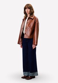 Jonge vrouw in een bruine leren jas, witte top, wijdlopende donkerblauwe jeans met cuffs en bruine loafers, staand tegen een witte achtergrond.