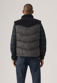 Chaleco acolchado con parte superior negra y sección inferior gris, con una textura acolchada y cuello alto, combinado con un suéter verde con patrón.