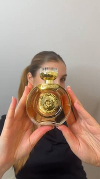 Une personne tient un flacon de parfum rond en verre avec des détails décoratifs dorés et un liquide de couleur ambre à l’intérieur.