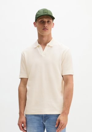 Junger Mann trägt ein cremefarbenes kurzärmeliges Poloshirt, blaue Jeans und eine olivgrüne Baseballkappe, steht vor einem weißen Hintergrund.