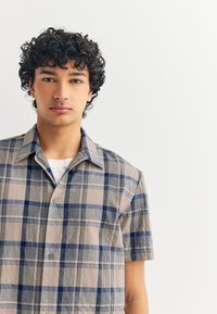 Short-sleeve, button-up shirt in a plaid pattern with beige, navy, and gray colors. Soft texture, collared design, and two buttons visible.