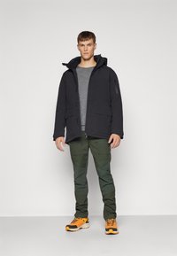 Czarna kapturowa kurtka z przednimi kieszeniami, szara koszulka pod spodem, zielone spodnie z kieszeniami oraz pomarańczowe buty sportowe z czarnymi akcentami.