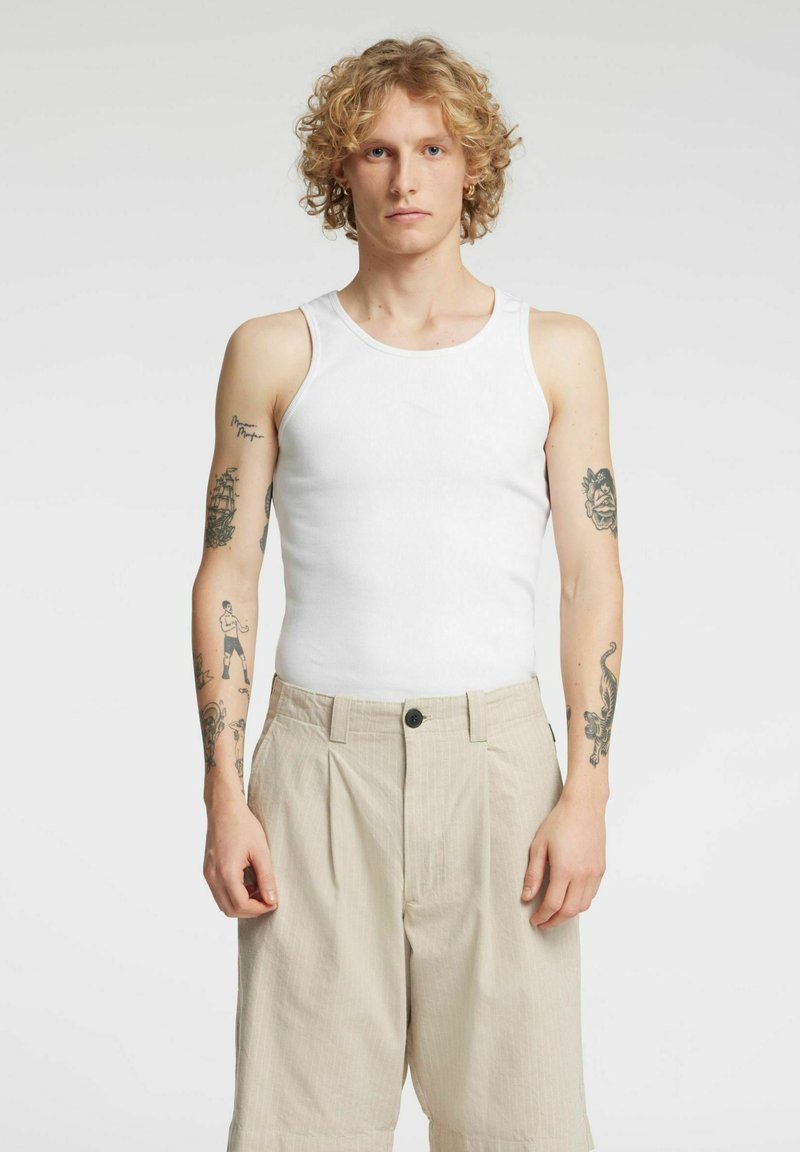 Biały bawełniany tank top z szerokimi ramiączkami, noszony z beżowymi plisowanymi szortami. Widoczne tatuaże na ramionach, jednolite tło.