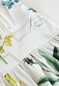 Lähivõte valgest kangast, millel on prinditud kollased ja sinised lilled, rohelised lehed ning käsitsi kirjutatud tekst "Laven".