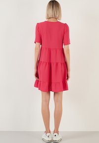 Růžové šaty s volány, krátkými rukávy, kulatým výstřihem a nabíraným lemem. Nošeny s bílými teniskami. Hladký materiál.