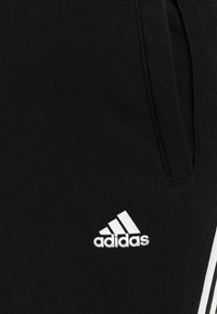 Pantalones de chándal negros de algodón con franjas laterales blancas y un pequeño logo de Adidas blanco en la parte inferior de la pierna. Son visibles dos bolsillos.