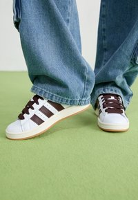 Zapatillas blancas de cuero con rayas marrones, cordones marrones y suela de goma, combinadas con unos pantalones de mezclilla azul holgados sobre una superficie verde.