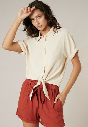 Femme aux cheveux ondulés foncés portant un haut crème à manches courtes boutonné et noué à la taille, ainsi qu'un short à cordon de serrage couleur rouille, posant avec une main levée.