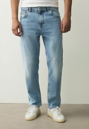 Mann steht in hellblauen Jeans, weißen Turnschuhen und einem weißen Hemd, gezeigt von der Taille bis zu den Füßen vor einfachem Hintergrund.