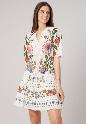 Femme portant une robe blanche à manches courtes avec une broderie florale colorée, taille cintrée et ourlet festonné, se tenant devant un fond uni.