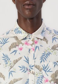 Camisa de manga corta en tejido ligero con un estampado de flores y hojas en tonos azules, rosas y beige, con cuello abotonado y bolsillo en el pecho.