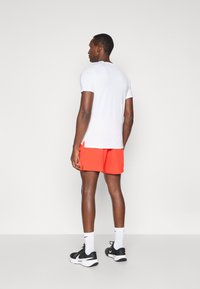 Άνδρας που στέκεται με την πλάτη, φορώντας λευκό μπλουζάκι, φωτεινές πορτοκαλί βερμούδες, λευκές κάλτσες και μαύρα και λευκά αθλητικά παπούτσια.