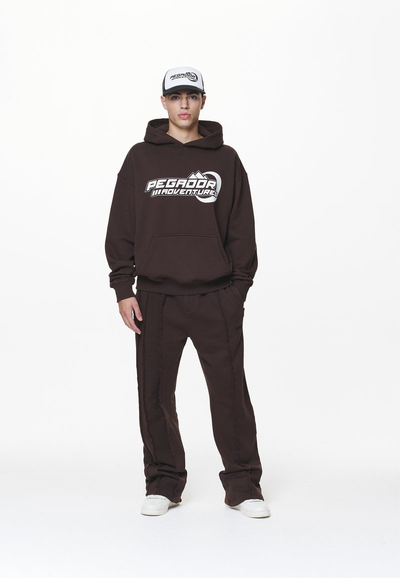 Brauner Hoodie mit "PEGADOR ADVENTURE" Grafik. Passende braune Sweatpants mit seitlichen Details. Weiße Sneakers und eine graue Mütze runden das Outfit ab.