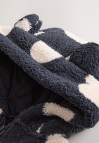 Veste à capuche en fausse fourrure avec des taches bleu marine et crème, présentant une texture pelucheuse et une doublure intérieure foncée contrastante. La capuche comprend de petites oreilles.