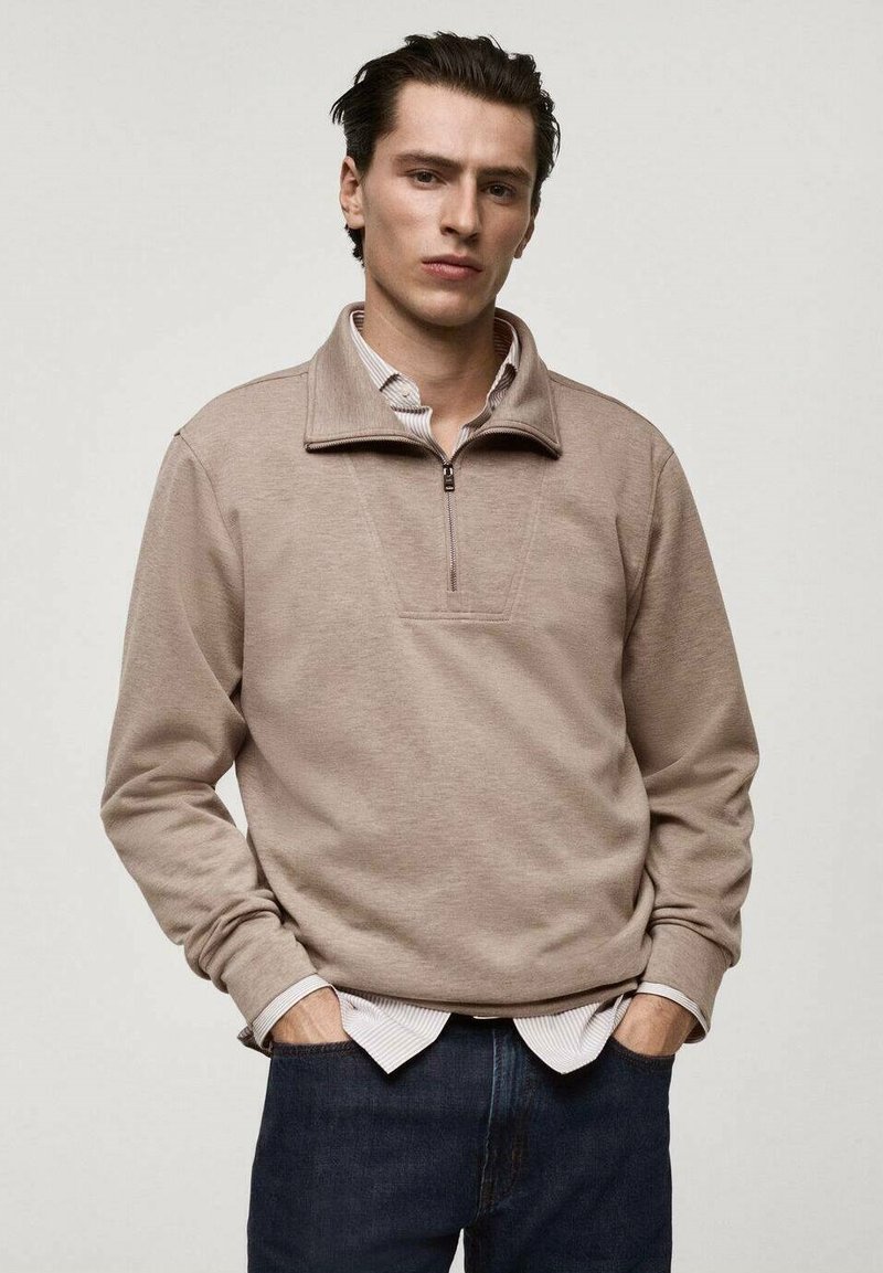 Pullover beige a mezza zip in tessuto morbido, con colletto alto e tasca frontale, indossato sopra una camicia a righe e abbinato a jeans scuri.