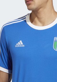 Blå sporttröja med vit krage och ärmdekorationer; har "adidas"-logotypen på vänster bröst och en färgglad lagbadge på höger sida.