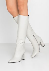 Botas de cuero blancas hasta la rodilla con punta afilada, tacón cuadrado grueso y detalle de cremallera lateral. Textura suave con suela negra contrastante.