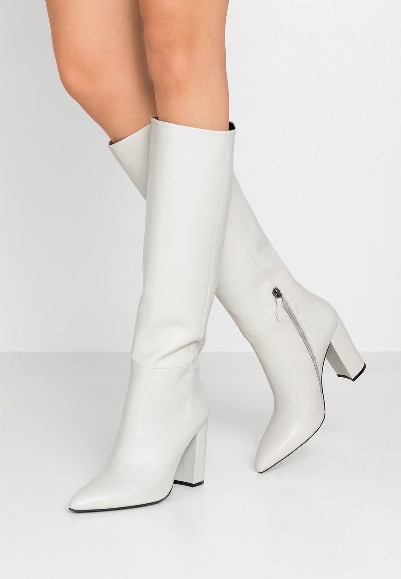 Botas de cuero blancas hasta la rodilla con punta afilada, tacón cuadrado grueso y detalle de cremallera lateral. Textura suave con suela negra contrastante.